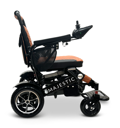 A side view of the MAJESTIC IQ-7000 Remote Controlled Electric Wheelchair showcases its sleek black design with a brown cushioned seat and backrest. The wheelchair features large rear wheels, smaller front wheels, a joystick control panel, and prominently displays the Majestic brand name on the frame.