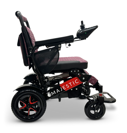 A side view of the MAJESTIC IQ-7000 Remote Controlled Electric Wheelchair showcases its sleek red and black color scheme, featuring an intelligent joystick control on the armrest, a cushioned seat, and large rear wheels for enhanced mobility. The brand name "MAJESTIC" is prominently displayed on the frame.