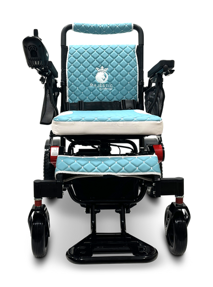 A front-facing view showcases the MAJESTIC IQ-7000 Remote Controlled Electric Wheelchair, featuring quilted light blue upholstery and a sleek black frame. Equipped with armrests, a joystick control panel, and a footrest, the wheelchair proudly displays the Majestic brand name embroidered on the backrest, highlighting its superior design and functionality.