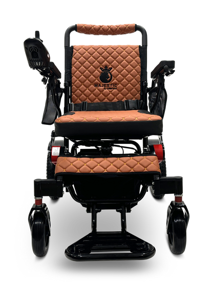 The MAJESTIC IQ-7000 Remote Controlled Electric Wheelchair boasts a lightweight design, featuring a quilted brown leather seat and backrest. It is equipped with joystick control on the left armrest and a sleek black metal frame that allows it to glide effortlessly on four wheels. The distinguished design is enhanced by the elegantly embroidered MAJESTIC logo.
