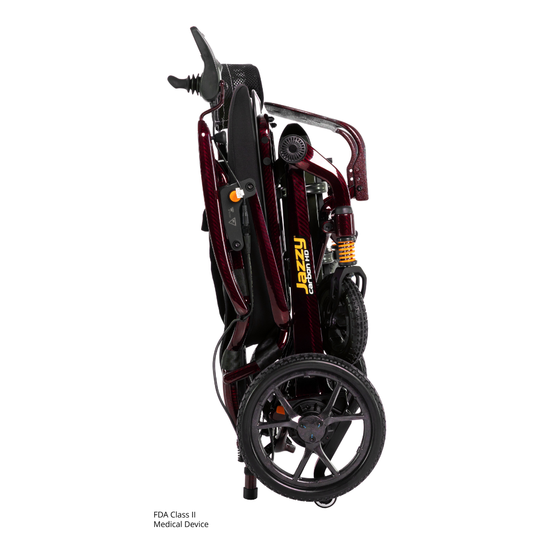 The Pride Jazzy Carbon HD Bariatric Portable Powerchair, shown here folded upright in burgundy, displays its wheels, seat, and handlebar with the Jazzy logo visible on the side.