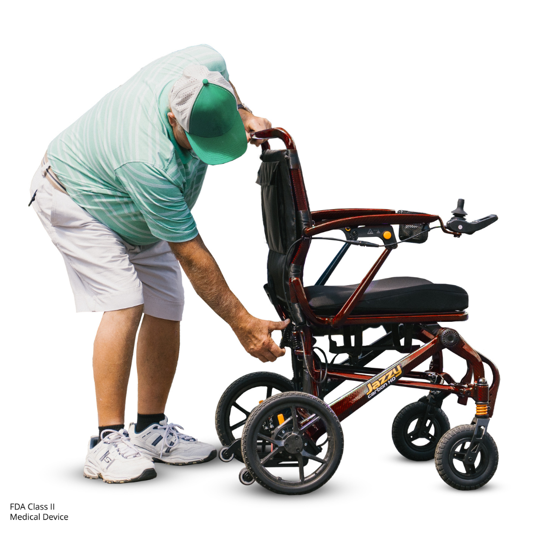 A man in a green-striped shirt, white shorts, and green cap adjusts a red Pride Jazzy Carbon HD Bariatric Portable Powerchair by turning a side knob. He is bending over against a white background.