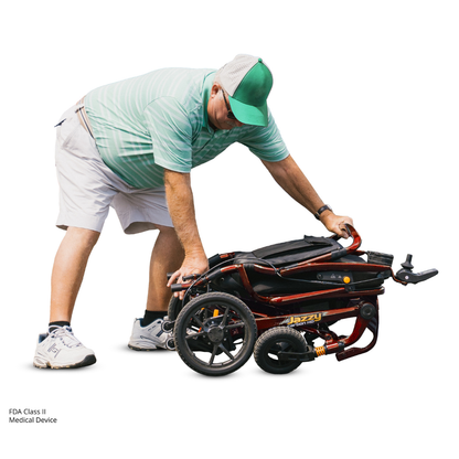 A man in a green cap and striped polo shirt bends over to fold or unfold a red Pride Jazzy Carbon HD Bariatric Portable Powerchair, with one knee slightly bent and hands gripping the frame.