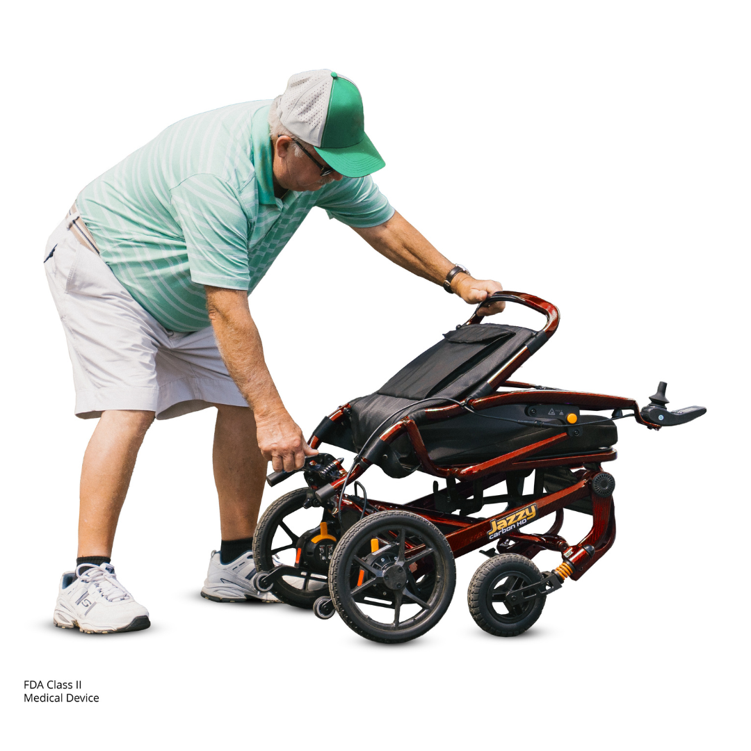 An older man in a green cap, striped polo, and shorts bends down to fold a red Pride Jazzy Carbon HD Bariatric Portable Powerchair, carefully adjusting the partially collapsed travel chair for transport.