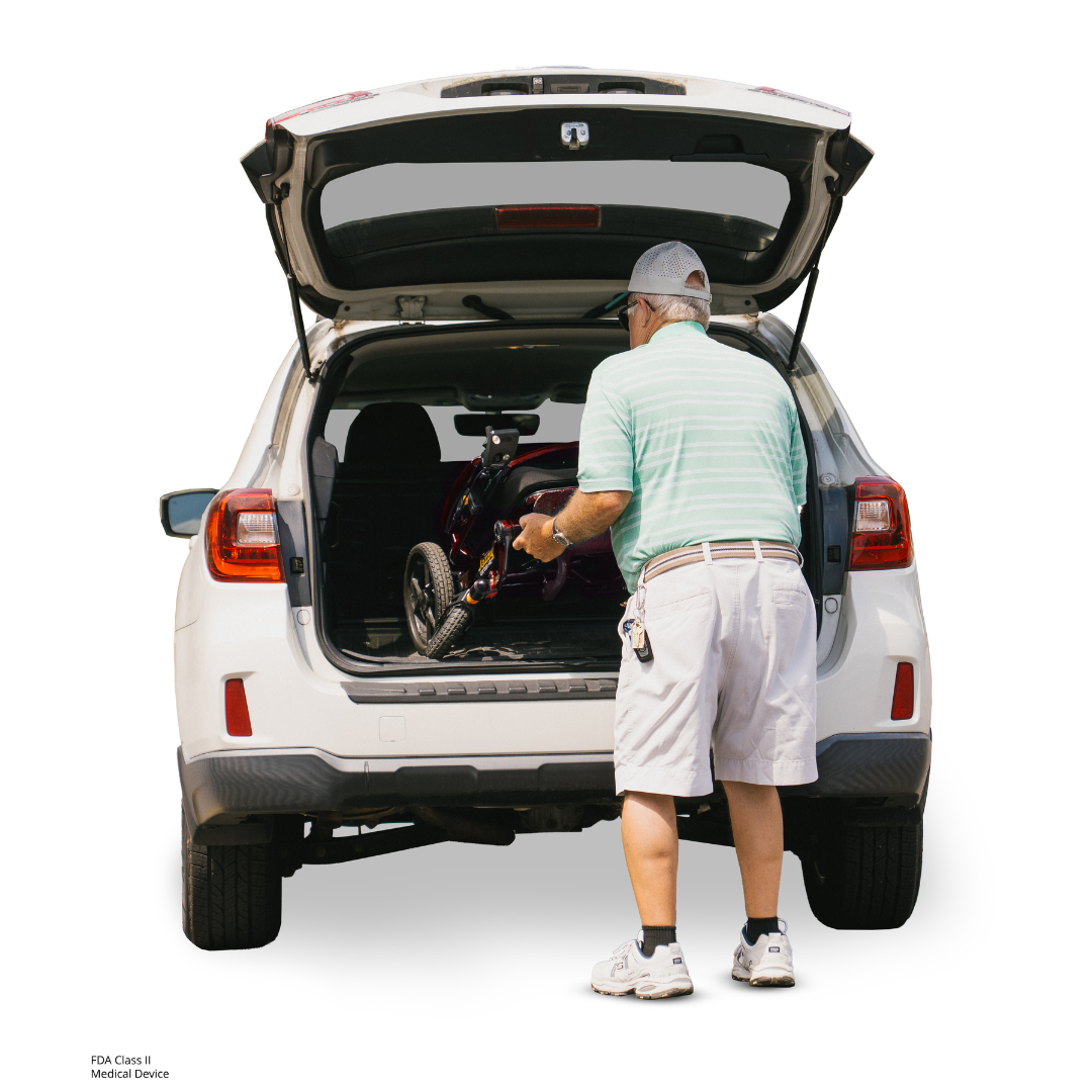 An older man in a cap, striped shirt, and shorts loads a folded Pride Jazzy Carbon HD Bariatric Portable Powerchair into the trunk of a white SUV with its rear hatch open.
