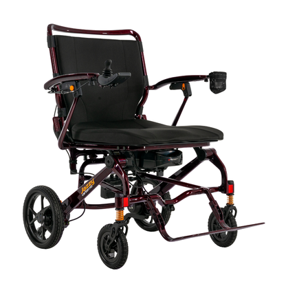 The Pride Jazzy Carbon HD Bariatric Portable Powerchair is a lightweight electric wheelchair with a black cushioned seat, red metallic frame, armrests, joystick control, four wheels, and a drink holder on one armrest.