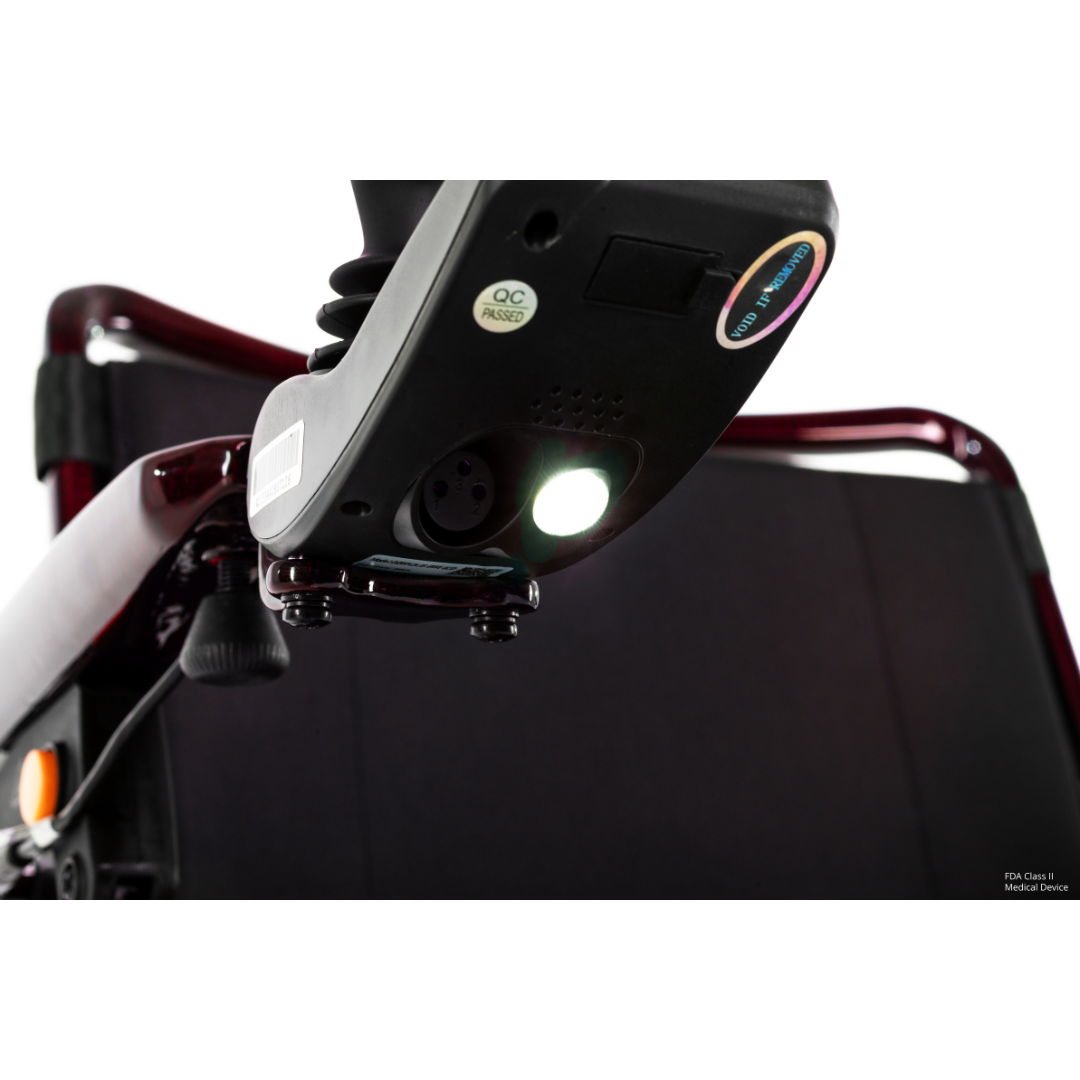 Close-up of the underside of the Pride Jazzy Carbon HD Bariatric Portable Powerchair’s control panel, featuring a glowing white LED light and multiple ports, with part of the wheelchair frame visible in the background.