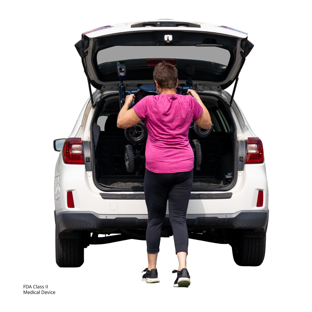 A person in a pink shirt and black pants loads a Jazzy® Carbon 27 Ultralite Portable Power Wheelchair into the trunk of a white SUV with the rear hatch open.