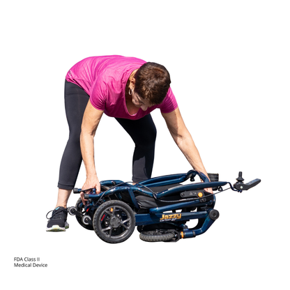 A woman in a pink shirt and black leggings lifts the folded Jazzy® Carbon 27 Ultralite Portable Power Wheelchair. The background is white with text reading "FDA Class II Medical Device" in the corner.