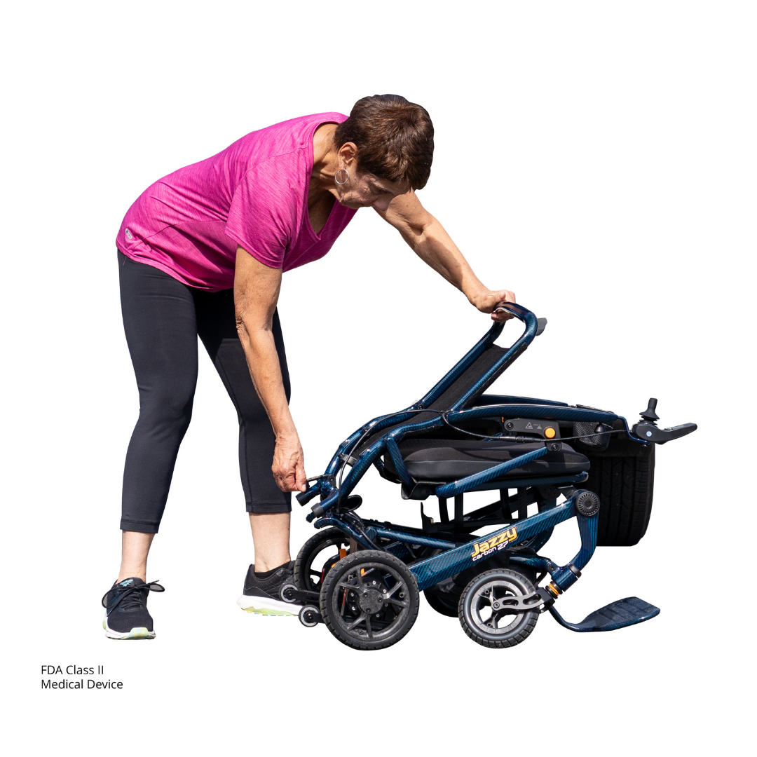 A woman in athletic wear bends down to fold or adjust a Jazzy® Carbon 27 Ultralite Portable Power Wheelchair against a white background.