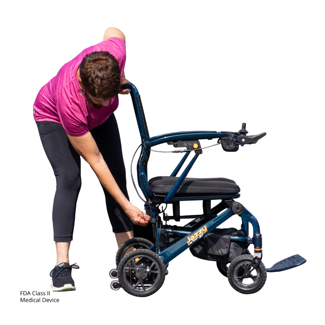 A woman in a pink shirt and black leggings adjusts a blue Jazzy® Carbon 27 Ultralite Portable Power Wheelchair set against a white background. "FDA Class II Medical Device" appears in text at the lower left corner.