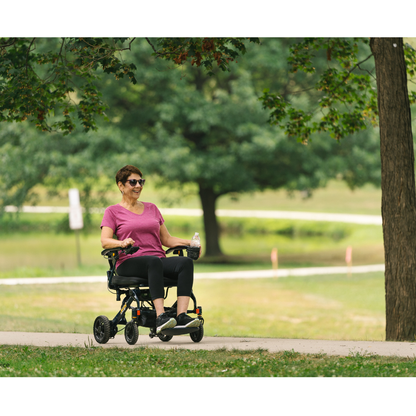 A woman in sunglasses and a pink shirt smiles while riding the Jazzy® Carbon 27 Ultralite Portable Power Wheelchair along a sunlit park path, surrounded by green grass and trees. She holds a water bottle as she enjoys her ride.