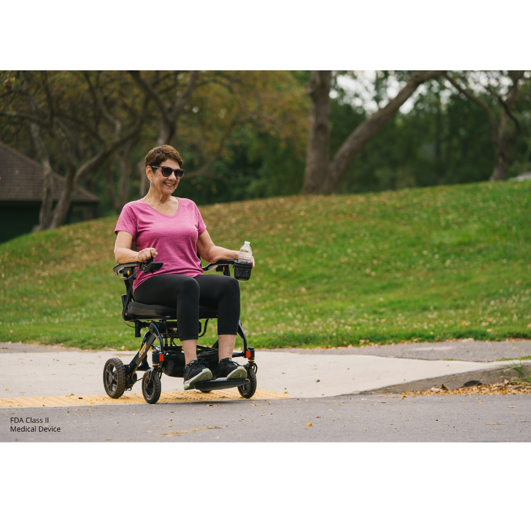 A woman in a pink shirt and sunglasses smiles while using a Jazzy® Carbon 27 Ultralite Portable Power Wheelchair outdoors on a paved path, holding a water bottle. Green grass and trees are in the background.