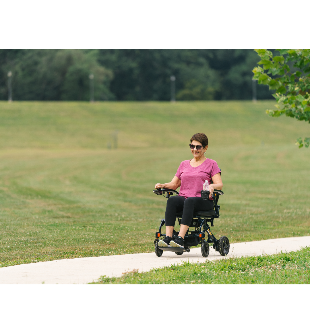 A woman in sunglasses and a pink shirt rides the Jazzy® Carbon 27 Ultralite Portable Power Wheelchair along a paved path in a grassy park on a sunny day.