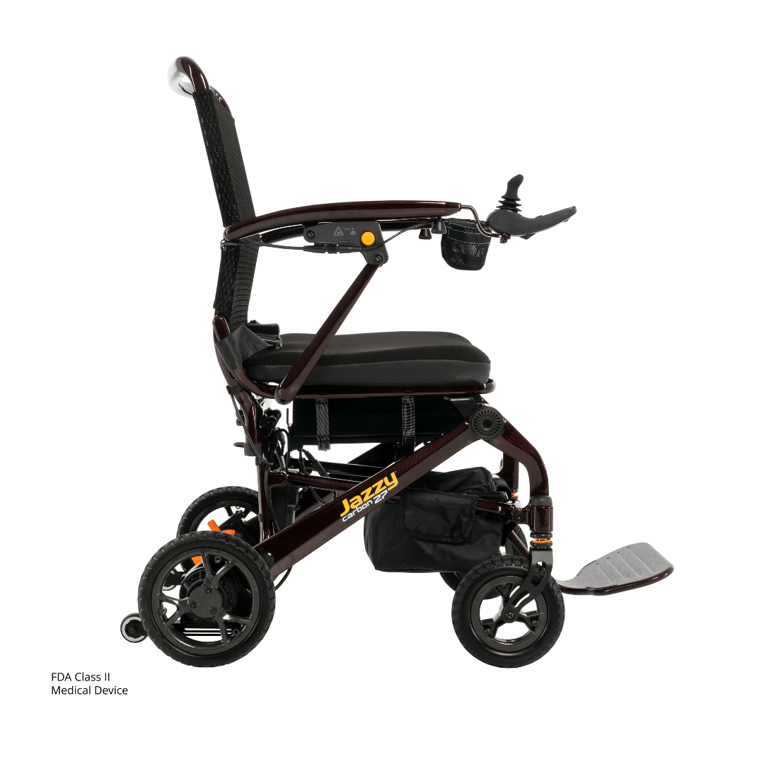 The Jazzy® Carbon 27 Ultralite Portable Power Wheelchair is lightweight, foldable, with a black cushioned seat, armrests, footrest, joystick control, four wheels, storage pouch, and a sleek design for convenience.