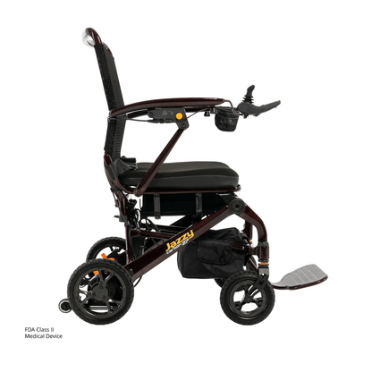 The Jazzy® Carbon 27 Ultralite Portable Power Wheelchair is lightweight, foldable, with a black cushioned seat, armrests, footrest, joystick control, four wheels, storage pouch, and a sleek design for convenience.