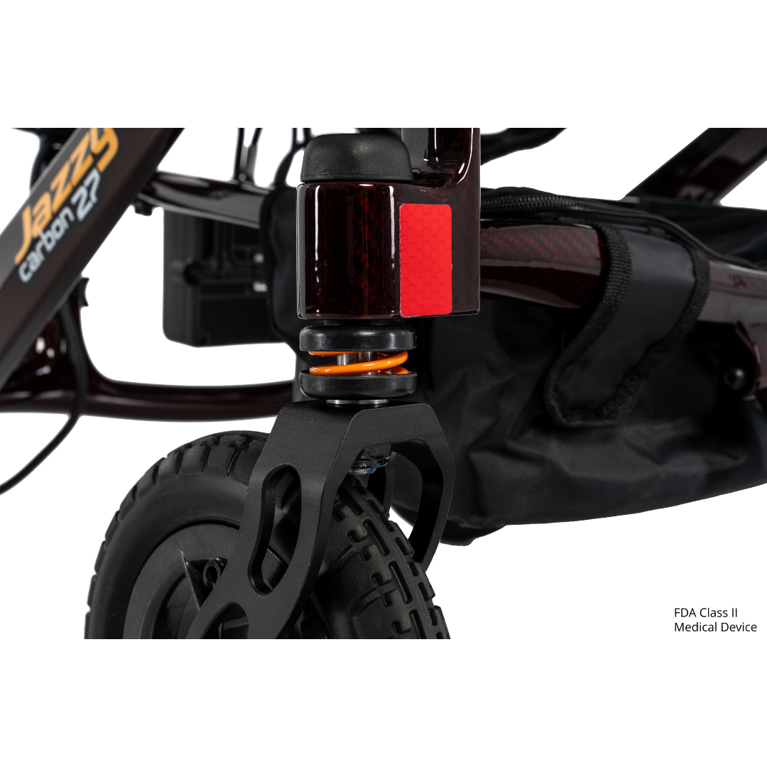 Close-up of the front wheel and suspension of the Jazzy® Carbon 27 Ultralite Portable Power Wheelchair, featuring a black tire, orange shock absorber, red reflector, partial storage bag, and "FDA Class II Medical Device" text in the lower right corner.