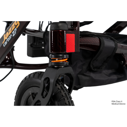 Close-up of the front wheel and suspension of the Jazzy® Carbon 27 Ultralite Portable Power Wheelchair, featuring a black tire, orange shock absorber, red reflector, partial storage bag, and "FDA Class II Medical Device" text in the lower right corner.
