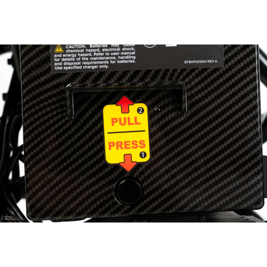 Close-up of a Pride Jazzy® Carbon 27X Portable Power Wheelchair, showcasing its black carbon fiber frame. A yellow sticker instructs: PRESS (1) with a downward arrow, PULL (2) with an upward arrow. A black button is below the sticker.