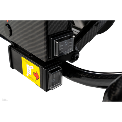 Close-up of two black switches with white markings on the Pride Jazzy® Carbon 27X Portable Power Wheelchair, beside a yellow label with a red arrow and battery icons. The device has a carbon fiber texture, typical of ultra-lightweight mobility chairs.