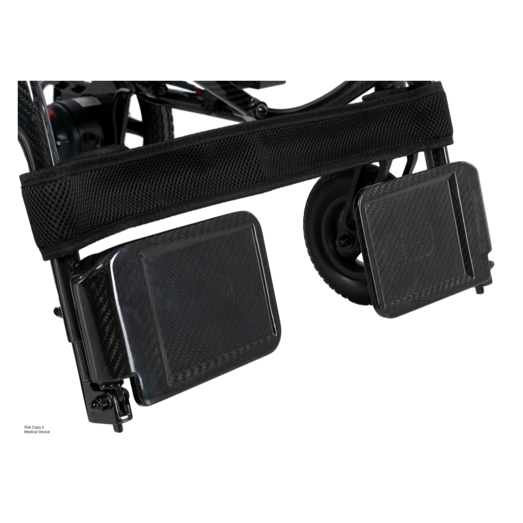 Close-up of two black rectangular footplates with a mesh strap above them, attached to the frame and small wheels of the Pride Jazzy® Carbon 27X Portable Power Wheelchair.