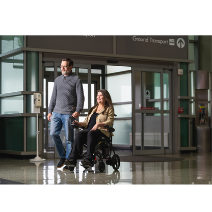 A man walks beside a woman using the Pride Jazzy® Carbon 27X Portable Power Wheelchair near airport automatic doors, beneath ground transport signs. The bright terminal features glass walls and polished floors.