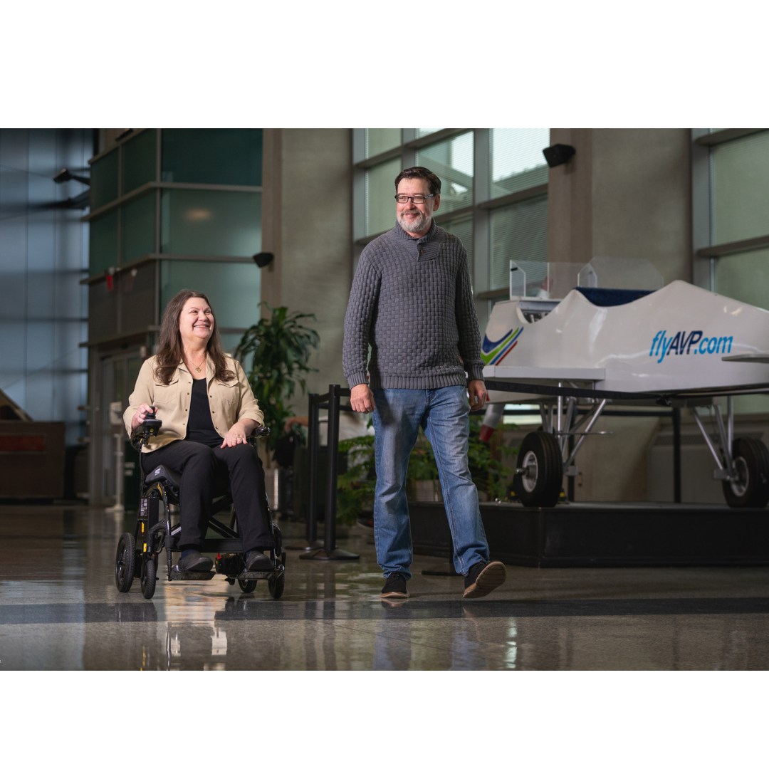 A woman in a Pride Jazzy® Carbon 27X Portable Power Wheelchair smiles at a man walking beside her indoors, with a small display aircraft and flyAVP.com visible in the background.