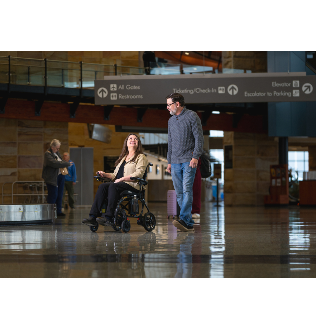 A woman smiles in a Pride Jazzy® Carbon 27X Portable Power Wheelchair while a man walks beside her through an airport terminal with signs for gates, restrooms, ticketing, check-in, elevators, and parking overhead.
