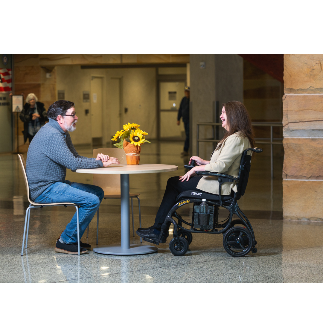 A man and a woman, one seated in a Pride Jazzy® Carbon 27X Portable Power Wheelchair, talk and smile across a round table with sunflowers in a bright, spacious room with stone walls.