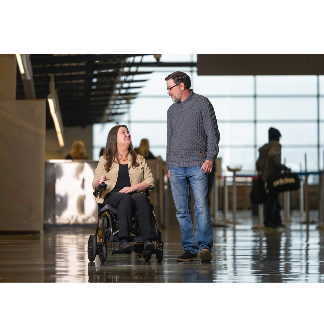 A woman in a Pride Jazzy® Carbon 27X Portable Power Wheelchair and a man walk side by side, smiling at each other in a bright indoor space with large windows and several people in the background.