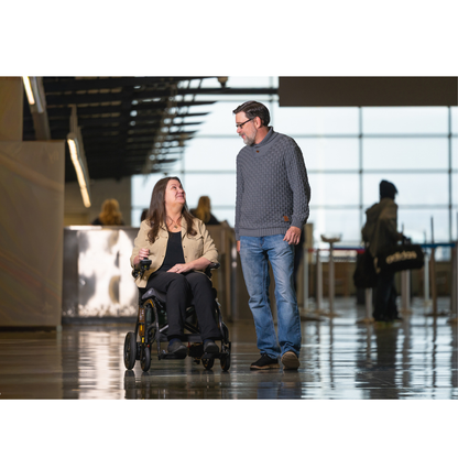 A woman in a Pride Jazzy® Carbon 27X Portable Power Wheelchair and a man walk side by side, smiling at each other in a bright indoor space with large windows and several people in the background.