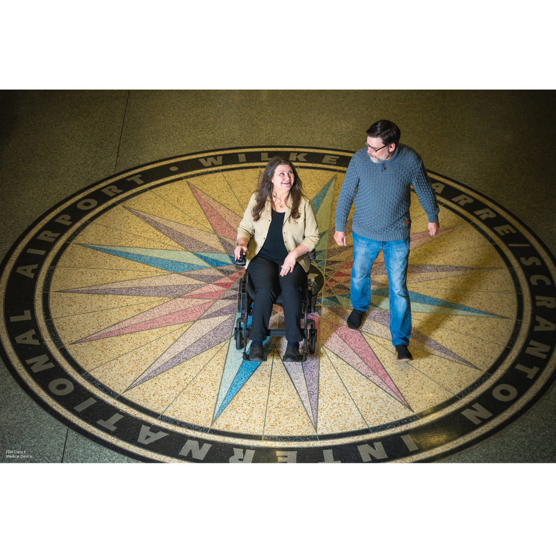 A woman using the Pride Jazzy® Carbon 27X Portable Power Wheelchair and a man stand smiling on a vibrant compass at Wilkes-Barre/Scranton International Airport, showcasing effortless travel with this modern mobility chair.