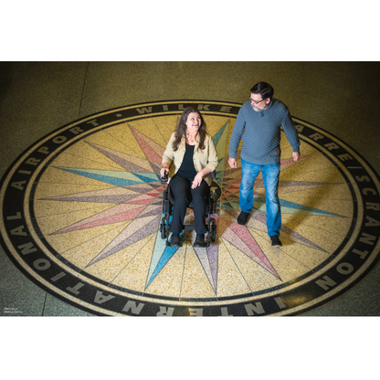 A woman using the Pride Jazzy® Carbon 27X Portable Power Wheelchair and a man stand smiling on a vibrant compass at Wilkes-Barre/Scranton International Airport, showcasing effortless travel with this modern mobility chair.