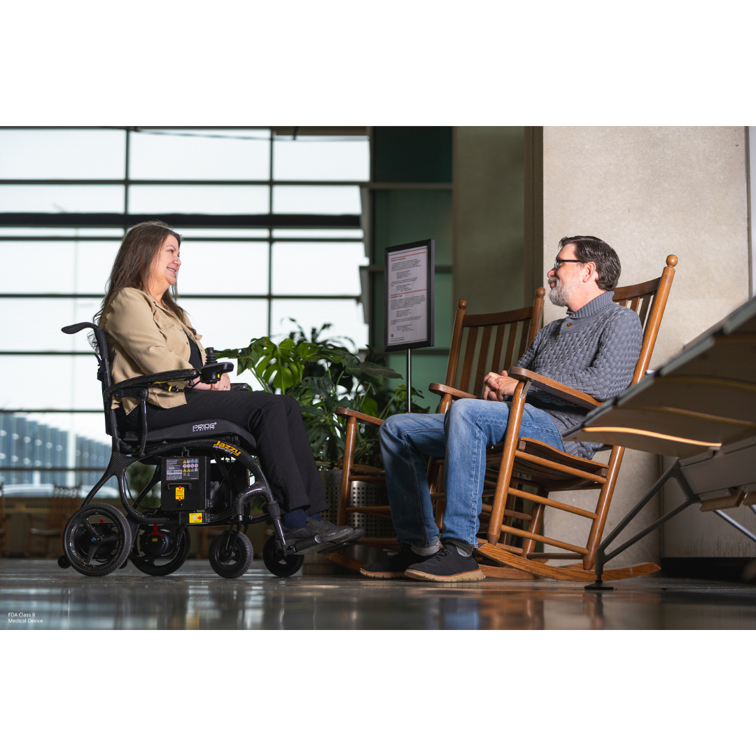 A woman using the Pride Jazzy® Carbon 27X Portable Power Wheelchair and a man in a rocking chair sit indoors, conversing face-to-face, with large windows and greenery visible behind them.