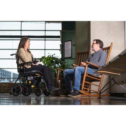 A woman using the Pride Jazzy® Carbon 27X Portable Power Wheelchair and a man in a rocking chair sit indoors, conversing face-to-face, with large windows and greenery visible behind them.