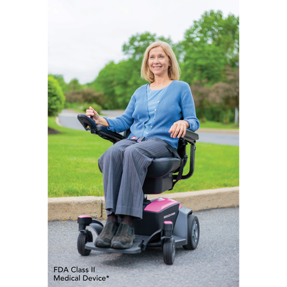 A smiling woman sits outdoors on a paved path in a Pride Mobility Go Chair® Portable Powerchair, wearing a blue cardigan, gray pants, and black shoes. Green grass, trees, and bushes are visible in the background.