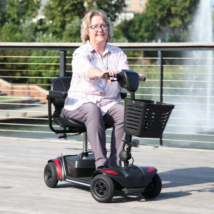 A woman smiles as she enjoys the intuitive controls of the Journey Adventure Scooter - 4 Wheel Portable Mobility Scooter on a wooden deck by the water. Clad in a light patterned blouse and gray pants, she appreciates its stability amid trees and a fountain enhancing the serene setting.