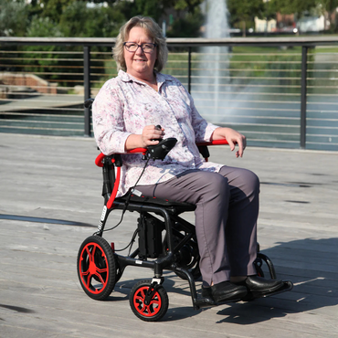 A woman in a floral blouse and gray pants sits confidently on her Journey Air Elite Folding Carbon Fiber Powerchair, weighing just 29 lbs, on a wooden deck by the lake. She smiles while holding her joystick control, with a fountain and greenery visible behind her.