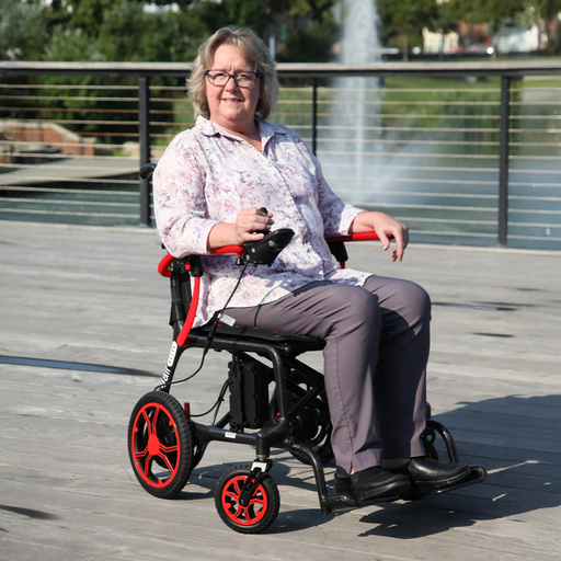 A woman in a floral blouse and gray pants sits in a Journey Air Elite Folding Lightweight Powerchair on a wooden deck, with a pond fountain and greenery behind her. Smiling at the camera, she highlights the chairs effortless indoor navigation thanks to its mere 29 lb weight.