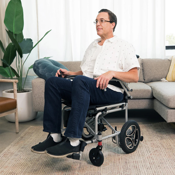 A person in a patterned white shirt and jeans sits in the Journey Air Lightweight Folding Powerchair, only 37 lbs, within a cozy living room with a sofa, green plant, and large window. The dual-drive system lets them maneuver easily as they look forward in the well-lit space.