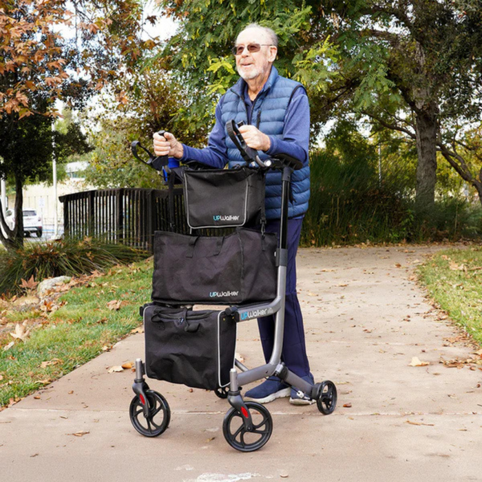 An elderly man in sunglasses and a blue vest uses the Journey UPWalker® EZ Lite Rollator along a tree-lined park path with scattered fallen leaves. This lightweight walker provides enhanced balance and includes attached storage bags.