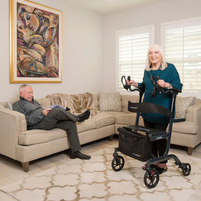 An elderly woman stands with a Journey UPWalker® Lite Upright Rolling Walker Rollator, smiling, while an elderly man reads on the sofa. The lightweight 15 lb walker supports her upright posture in a room filled with modern furniture and a colorful abstract painting, creating a lively ambiance.