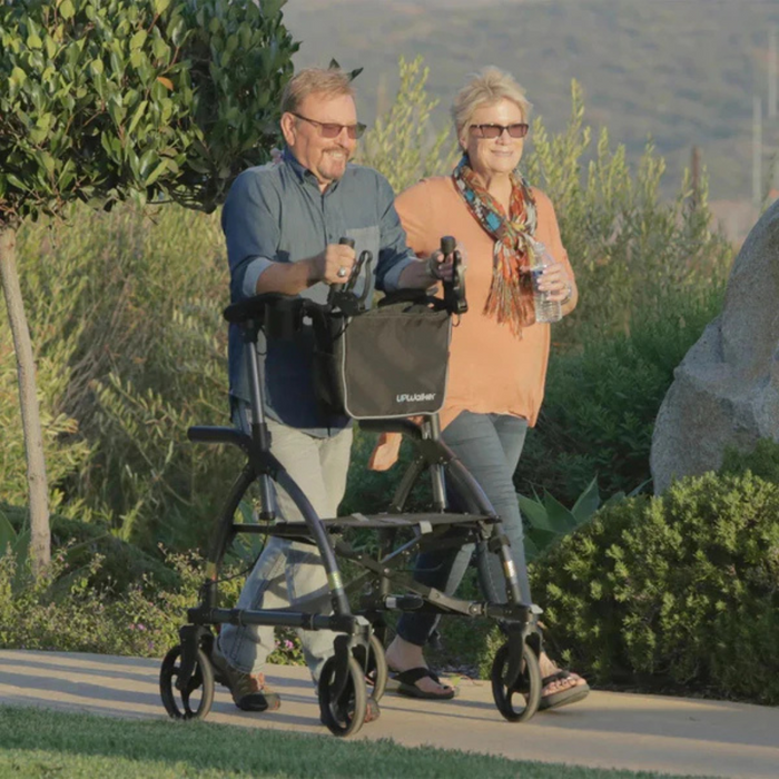 A man with a Journey UPWalker® Original Upright Rolling Walker Rollator and a woman with a water bottle stroll on a scenic, paved path amid greenery and hills, both wearing sunglasses and smiling brightly as they enjoy their outing.