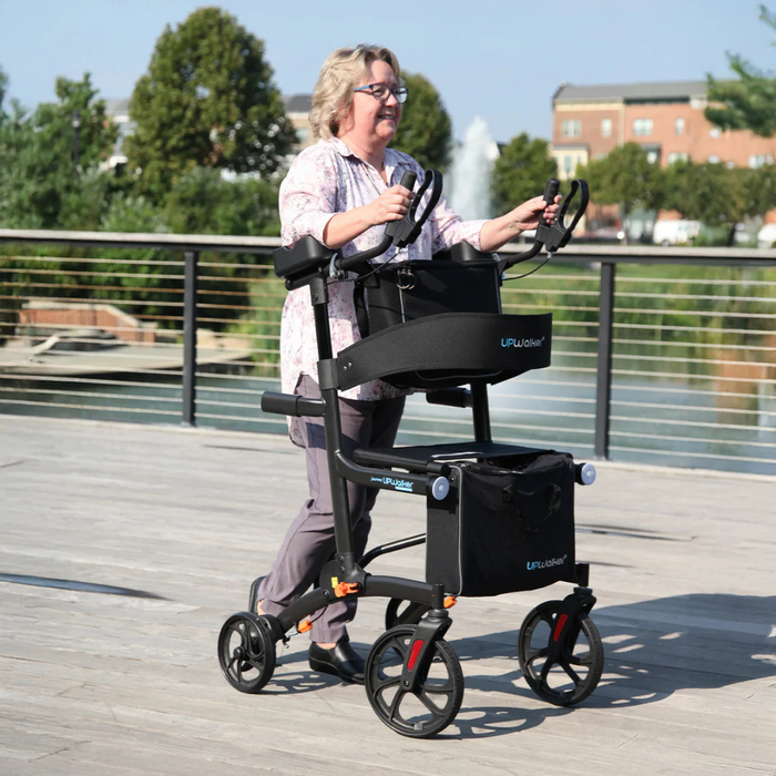 A woman uses a black Journey UPWalker Premium Lite V2 on a sunny wooden boardwalk, with trees and a building in view. The walker features four enhanced wheels and an attached storage bag.