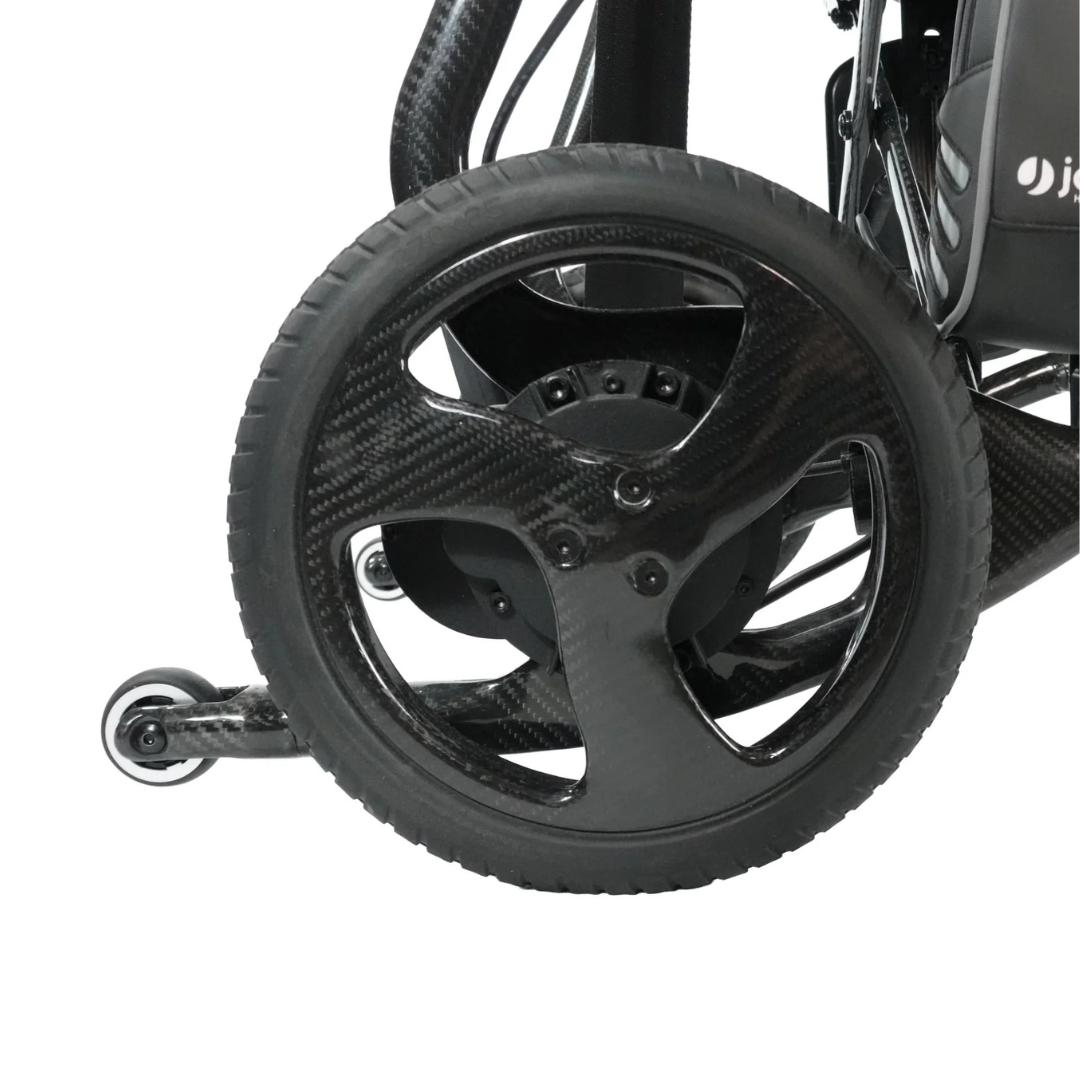 Close-up of the rear wheel of the Journey Air Elite Max Ultralite Carbon Fiber Travel Powerchair, showcasing its black three-spoke design and a small anti-tip wheel behind it against a white background.
