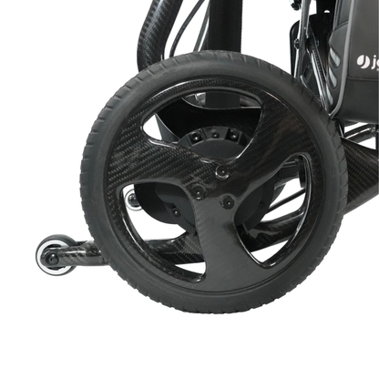 Close-up of the rear wheel of the Journey Air Elite Max Ultralite Carbon Fiber Travel Powerchair, showcasing its black three-spoke design and a small anti-tip wheel behind it against a white background.