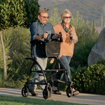 Two older adults walk outdoors on a path; the man uses the Journey UPWalker® Original Upright Rolling Walker Rollator as his mobility aid while the woman walks beside him holding a water bottle. Both are smiling and wearing sunglasses, with green bushes and hills in the background.