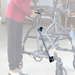 Indoors, a person in a red shirt and dark pants places a black and white walking cane onto a gray walker with wheels. The focus is on mobility assistance items, enhanced by Journey UPWalker Parts & Accessories for added ease.