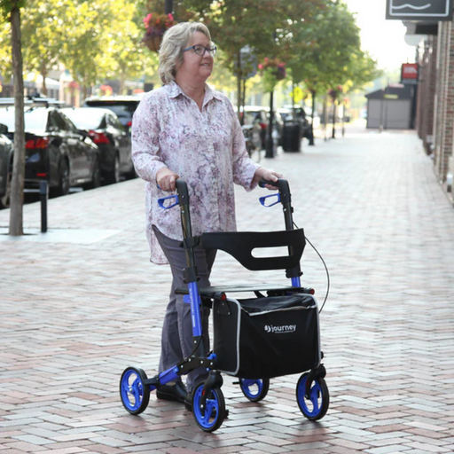 A woman with gray hair walks on a brick-paved sidewalk using her Journey UPWalker® Breeze Rollator Walker. In floral shirt and pants, she maintains improved posture as she moves. Trees and parked cars line the street in the background.