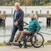 A man walks beside a woman in a Journey So Lite® C2 Ultra Lightweight Wheelchair along the waterfront. Both are smiling and enjoying the outdoor scenery with a dock and water in the background.